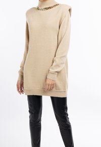 Pull en maille beige avec un col montant et un détail chaîne dorée, doté de manches longues et d'une coupe décontractée. Porté sur un pantalon en cuir noir.