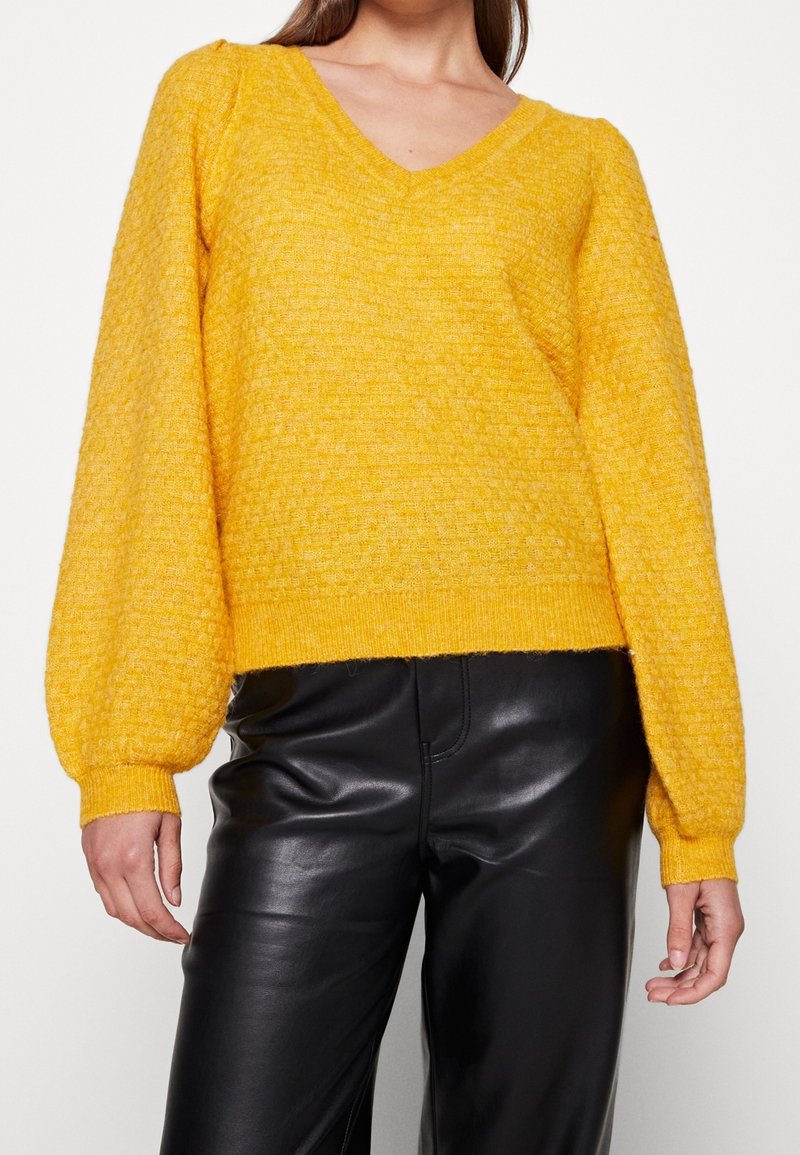 Femme portant un pull texturé jaune à col en V avec des manches ballon, assorti à un pantalon noir en similicuir.