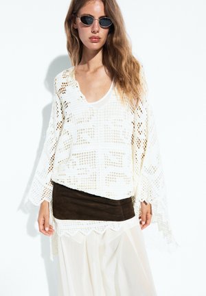 Femme aux longs cheveux et lunettes de soleil portant un top en dentelle blanche, une jupe blanche et une large ceinture marron foncé sur un fond blanc uni.