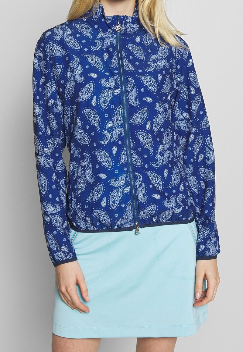 Modrá zipová bunda s paisley vzorem v bílé barvě, vyrobená z lehké tkaniny se stojáčkovým límcem a elastickým lemem.