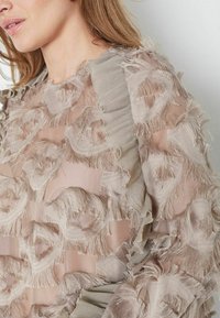 Průhledná blůza s vrstvenou, třepenou látkou v béžových odstínech. Obsahuje texturované květinové vzory a volánkové detaily podél okrajů.