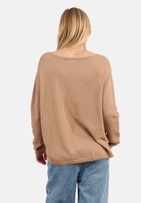Donna con lunghi capelli biondi, che indossa un maglione di maglia beige largo e Jeans blu, girata di spalle su uno sfondo bianco semplice.