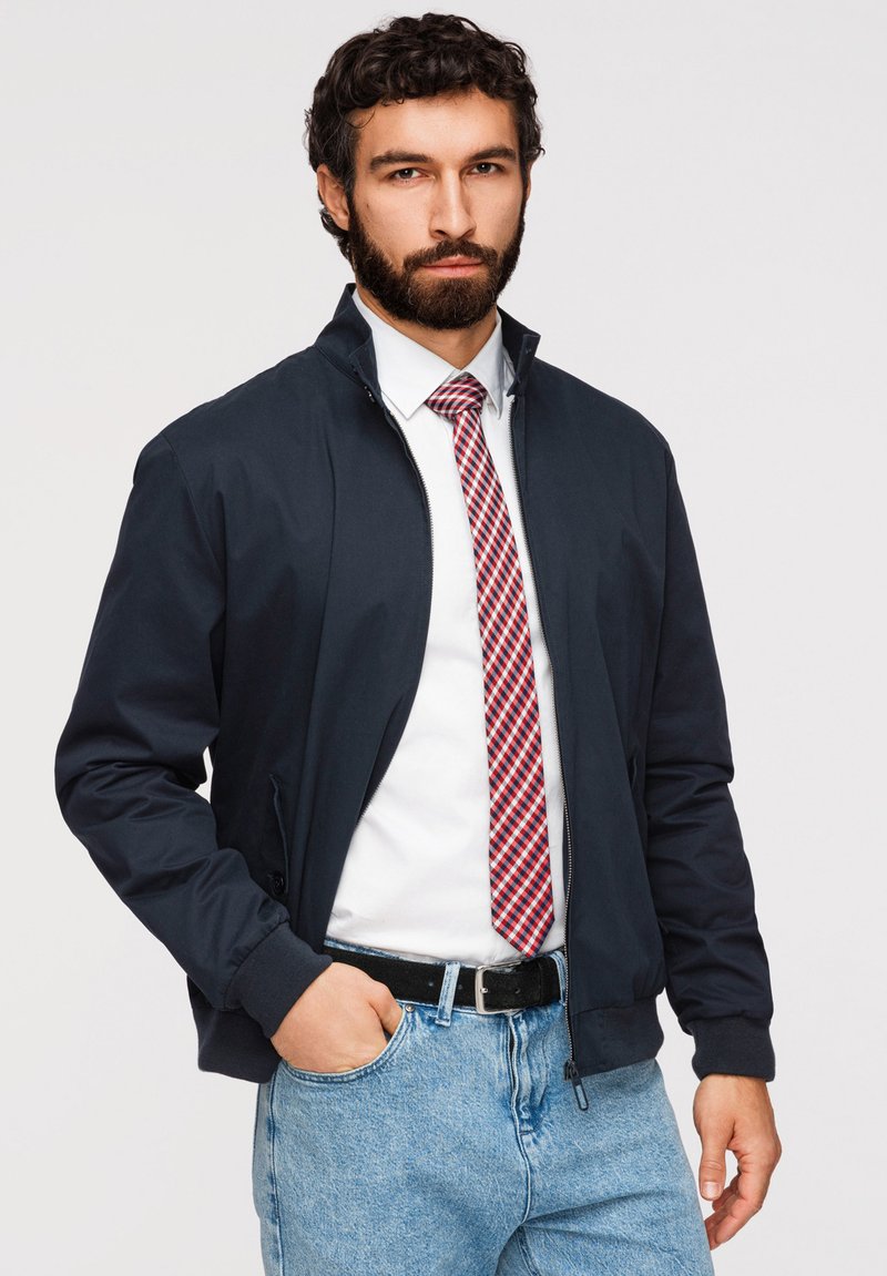 Homme barbu portant une veste bleu marine, une chemise blanche avec une cravate rouge à motifs, un jean bleu, une main dans la poche, debout devant un fond uni.