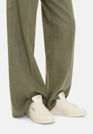 Osoba w oliwkowych spodniach o szerokich nogawkach nosząca kremowo-białe niskie sneakersy Reebok z sznurowadłami i złotymi detalami logo na białym tle.