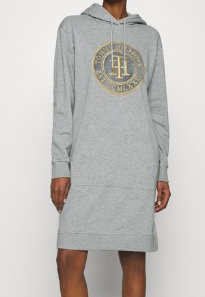 Szara sukienka z kapturem i kieszenią z przodu oraz złotym logo Tommy Hilfiger na piersi, noszona przez osobę o ciemnej karnacji.