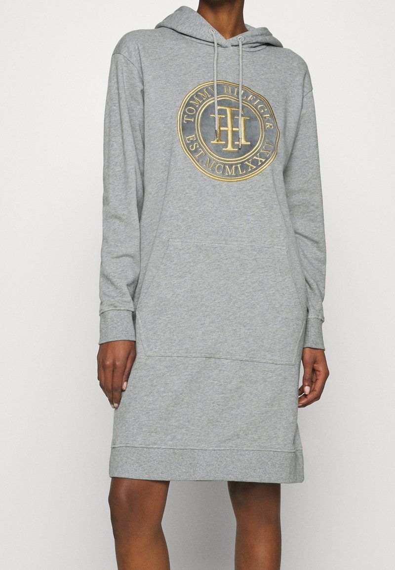 Robe sweat à capuche grise avec poche avant et logo Tommy Hilfiger doré sur la poitrine, portée par une personne à la peau foncée.
