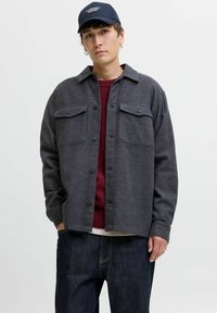 Camisa chaqueta de lana gris con dos bolsillos en el pecho, botones negros y cuello con solapa, usada sobre un suéter burdeos y jeans oscuros.