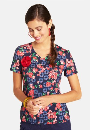 Young woman with braided hair wearing blue floral short-sleeve top, red earrings, and gold bangles, smiling with eyes closed and hands clasped.