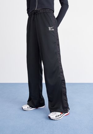 Person iført sorte Nike Air træningsbukser og hvide sportssko står på en blå gulv mod en ensartet hvid baggrund.