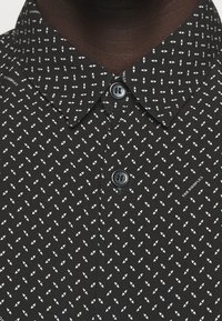 Schwarzes Hemd mit kleinen weißen geometrischen Mustern und Kragen, getragen von einer dunkelhäutigen Person, zwei Knöpfe geschlossen gezeigt.