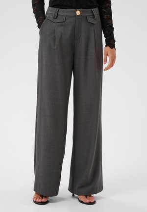 Femme portant un pantalon gris taille haute à jambes larges avec des plis sur le devant, un bouton doré et des poches latérales, associé à des sandales noires et un top en dentelle.