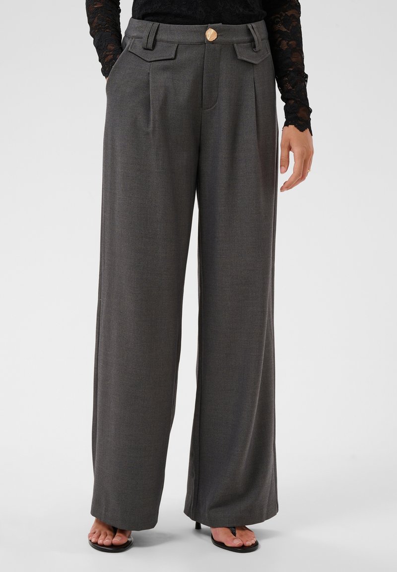 Femme portant un pantalon gris taille haute à jambes larges avec des plis sur le devant, un bouton doré et des poches latérales, associé à des sandales noires et un top en dentelle.