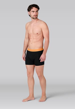 Mustad bokserid oranži vöökohtadega, valmistatud pehmest kangast, kohandatud sobivusega ja minimaalsete õmblustega. Modell seisab paljajalu.