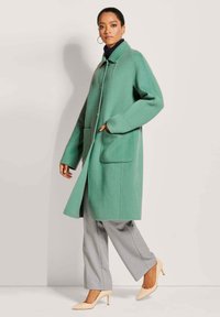 Groene wollen jas met grote opgestikte zakken, een kraag en knoopsluiting, gecombineerd met grijze broeken en naakte hoge hakken.