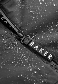 Schwarzer wasserdichter Stoff mit sichtbaren Wassertropfen, ausgestattet mit einem Reißverschluss und einem Etikett mit "BAKER" in Weiß auf einem strukturierten Band.