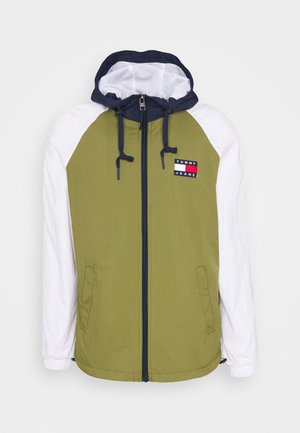 Giacca verde oliva con maniche bianche, cappuccio blu navy, zip frontale e tasche laterali. Presenta il logo "TOMMY JEANS" sul petto. Tessuto liscio.