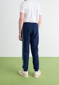 Marinblå träningsbyxor med elastisk midja och ljusblå sidostripes, i kombination med vita sneakers, stående på ett grönt golv.