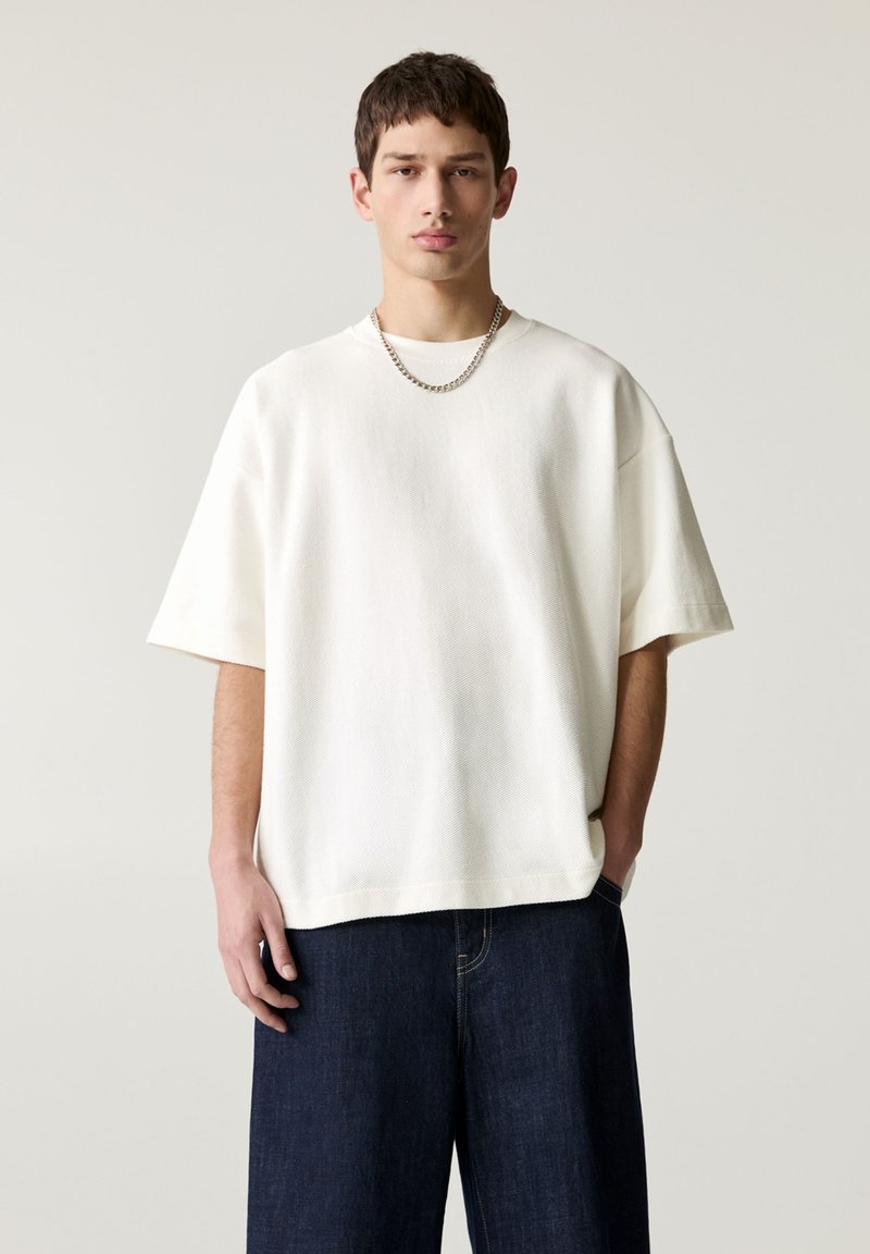 T-shirt blanc oversize en tissu texturé, avec des manches courtes et un col rond, associé à un jean en denim foncé ample.