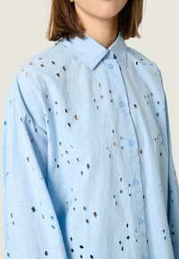 Lichtblauw shirt met een textuur stof en uitgesneden patronen, een knoopsluiting aan de voorkant en een klassieke kraag. Lange mouwen zijn zichtbaar.