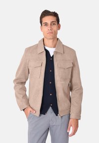 Giacca in misto lana beige con colletto, due tasche sul petto, chiusura zip e finitura testurizzata, indossata sopra una camicia blu navy con bottoni.