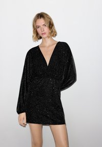 Frau mit kurzen blonden Haaren, die ein schwarzes, langärmliges, glitzerndes Mini-Kleid mit tiefem V-Ausschnitt vor einem weißen, schlichten Hintergrund trägt.