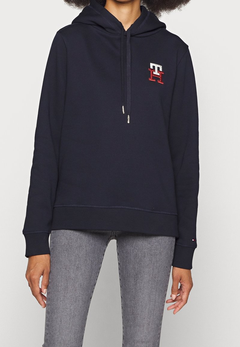 Personne portant un sweat à capuche bleu marine avec cordons, un logo "TH" rouge et blanc sur la poitrine, et un jean gris clair sur un fond uni.