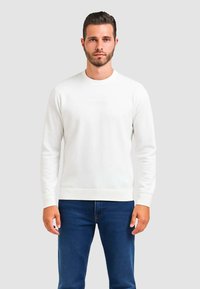 Felpa bianca con collo a giro, polsini e orlo a costine, caratterizzata da un logo in rilievo discreto sul petto. Indossata con jeans blu.