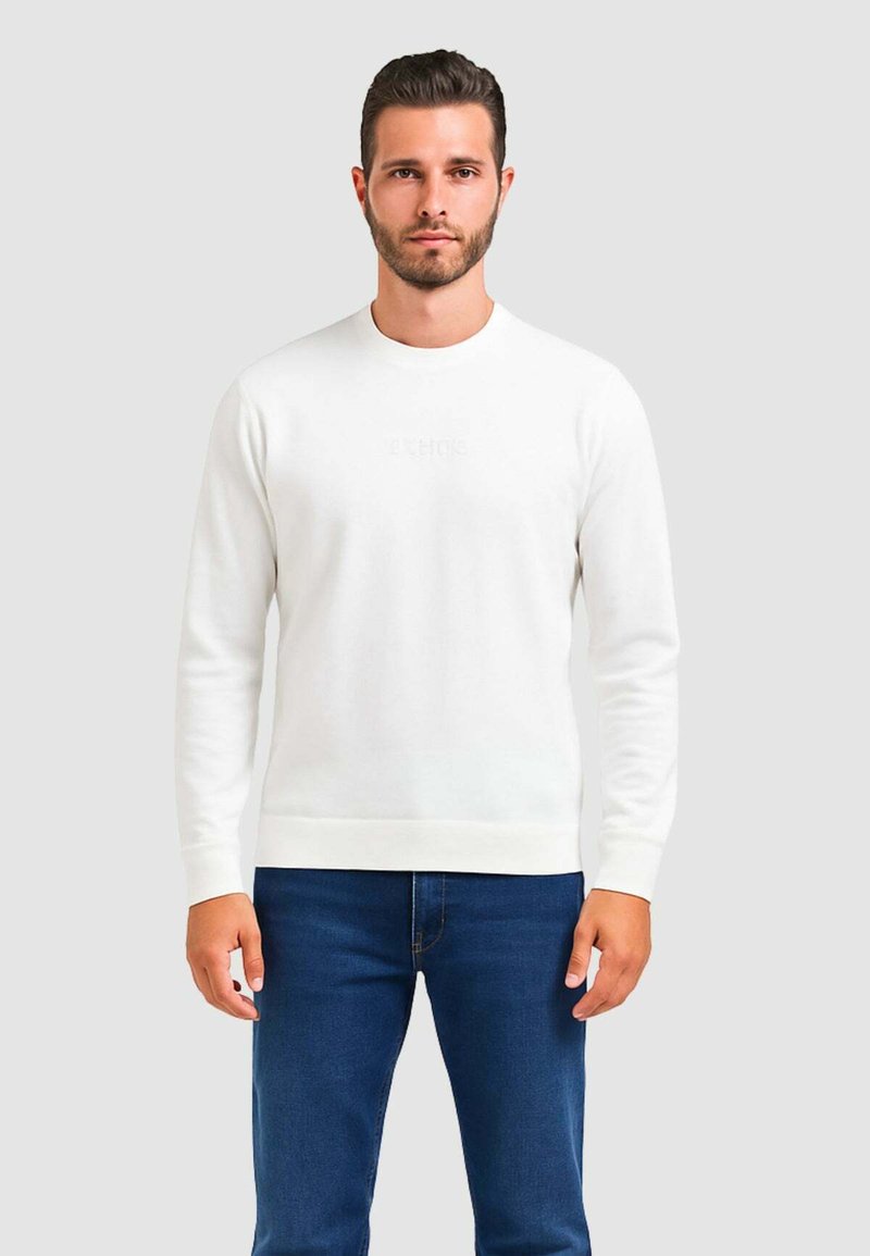 Felpa bianca con collo a giro, polsini e orlo a costine, caratterizzata da un logo in rilievo discreto sul petto. Indossata con jeans blu.