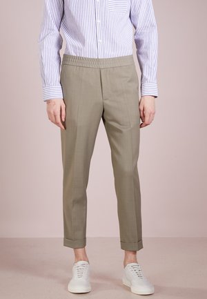 Hombre con pantalones beige de corte a medida con dobladillos, camisa de rayas abotonada y metida por dentro, y zapatillas blancas, de pie sobre un suelo beige.