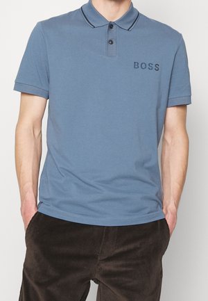 Man draagt een blauw korte mouw polo met twee zwarte knopen en het "BOSS"-logo op de borst, gecombineerd met donkerbruine corduroy broek.