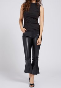 Vrouw in een mouwloos zwart topje, glanzende zwarte leren broek met uitlopende onderkanten en zwarte open teenschoenen, staande tegen een effen achtergrond.