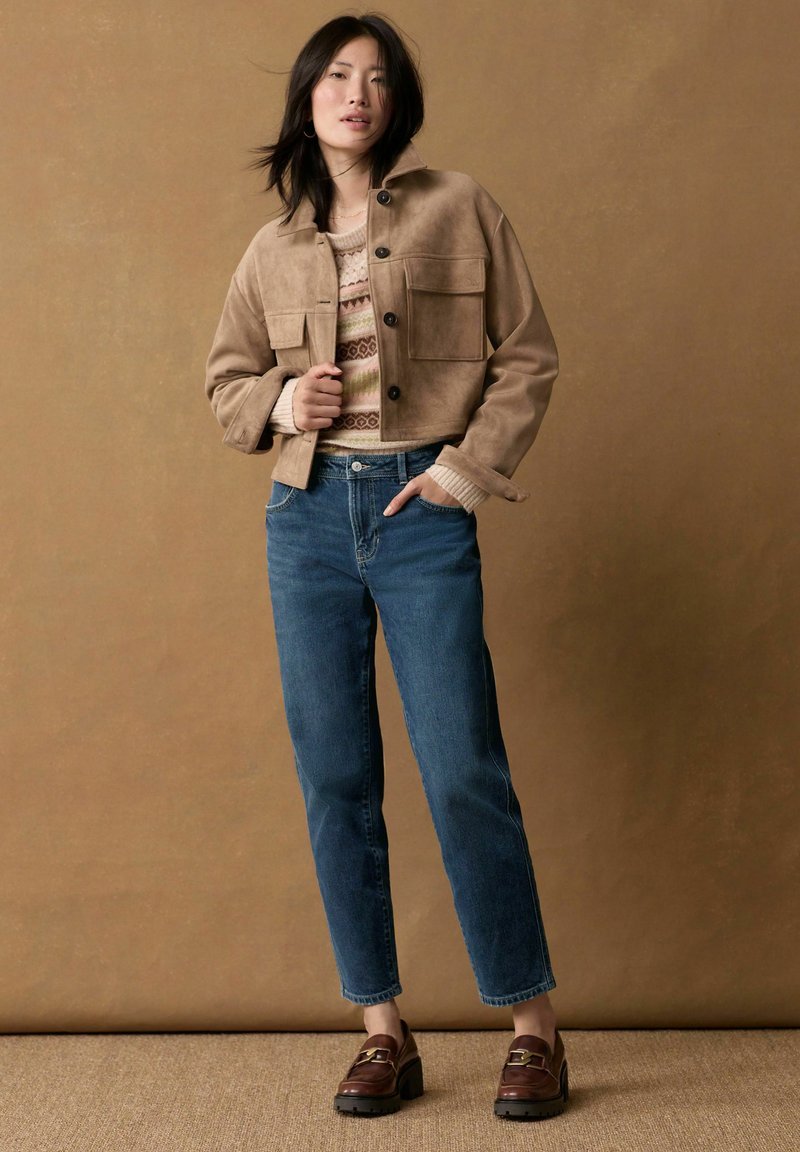 Tan cropped jack met twee voorzakken, gebreide trui in gedempte kleuren, blauwe denim jeans en bruine platform loafers op een gestructureerd oppervlak.