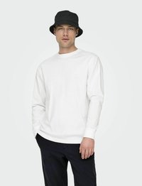 Witte katoenen blouse met lange mouwen en ronde hals, gecombineerd met een zwarte bucket hat. Model draagt een marineblauwe broek tegen een lichte achtergrond.