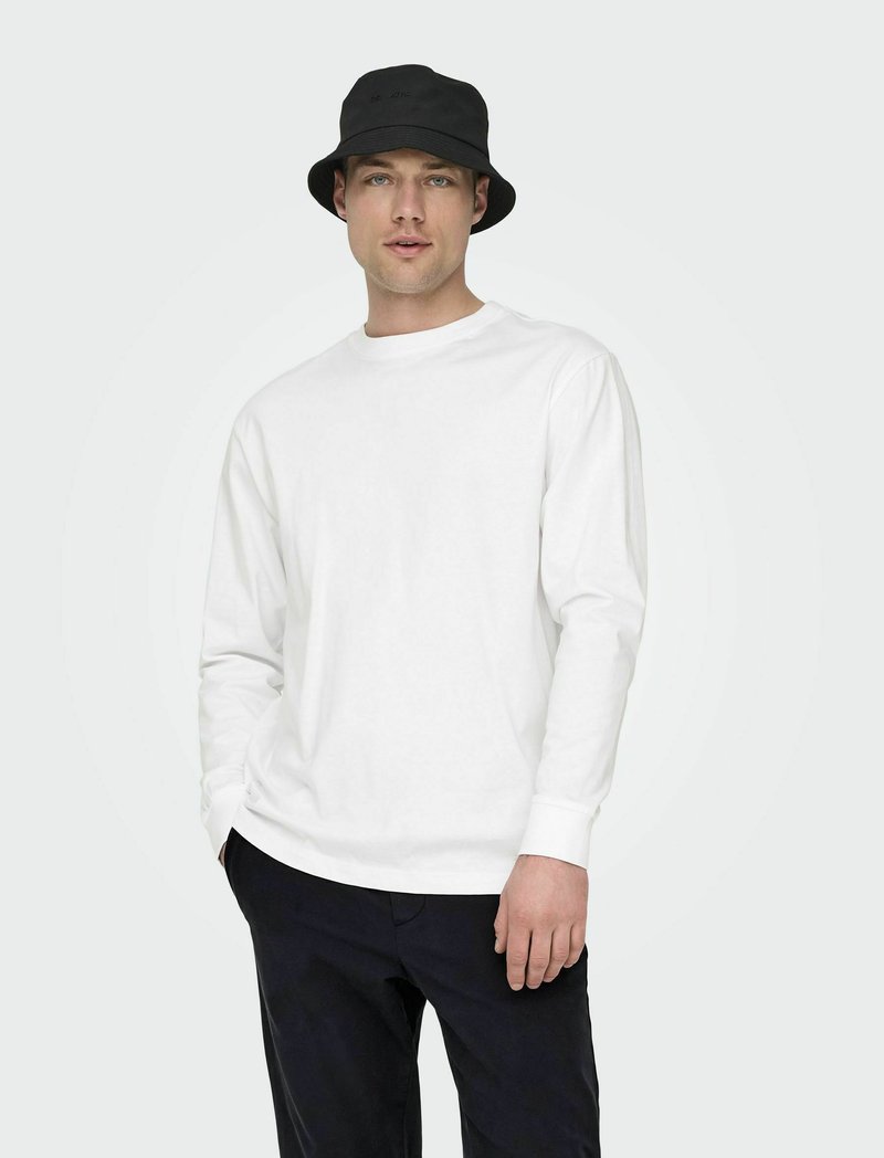 Witte katoenen blouse met lange mouwen en ronde hals, gecombineerd met een zwarte bucket hat. Model draagt een marineblauwe broek tegen een lichte achtergrond.