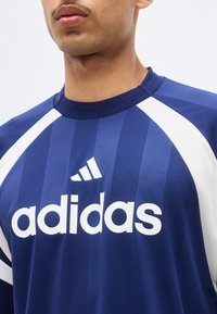 Άνδρας που φοράει μπλε αθλητική φανέλα Adidas με λευκές ρίγες και λογότυπο στο στήθος, εμφανιζόμενος από το λαιμό έως το μέσο του κορμού.