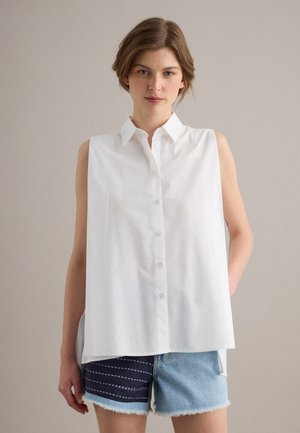 Joven mujer con cabello corto que lleva una camisa blanca sin mangas con botones y pantalones cortos de mezclilla bicolor, de pie frente a un fondo liso.