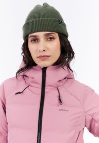 Růžová nafouknutá bunda s vysokým límcem a černým zipem, s tkaničkami. Na sobě má tmavě zelenou pletenou čepici.