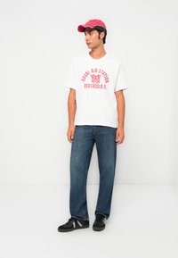 Young man wearing a red cap, white "Naval Air Station Pensacola" T-shirt, blue jeans, and black sneakers, standing against a white wall.
