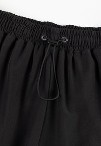 Czarne spodnie z elastycznym pasem i sznurkiem. Materiał wydaje się gładki i lekki, z plisami z przodu.