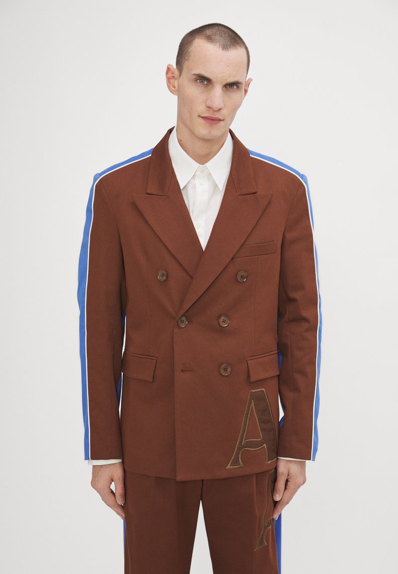 Blazer doppiopetto marrone con strisce laterali blu, dotato di bottoni in bronzo, una tasca sul petto e dettagli in lettera dorata sulla parte anteriore inferiore.
