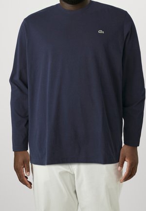 Uomo che indossa una camicia a maniche lunghe blu navy con un piccolo logo di coccodrillo verde sul petto e pantaloni bianchi, in piedi con le braccia rilassate.