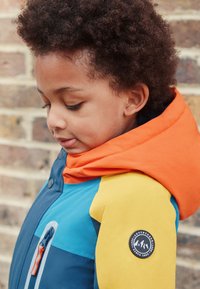 Kinderjack met twee kleuren, met blauwe, oranje en gele delen. Voorzien van een ritszak, ronde logopatch en een zachte capuchon.