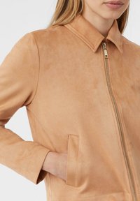Donna che indossa una giacca in suede marrone chiaro con zip frontale e mano nella tasca laterale, visibile dalle spalle alla vita.