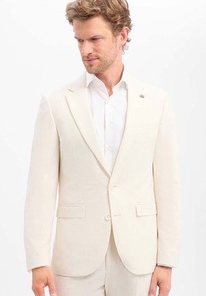 Man met lichtbruin haar, die een crèmekleurig colbert en een wit overhemd draagt, kijkt iets zijwaarts.