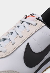 Λευκό αθλητικό παπούτσι Nike με μαύρο σήμα swoosh, λευκά κορδόνια και πορτοκαλί λογότυπο στη γλώσσα, απεικονίζεται σε κοντινή γωνιακή άποψη.