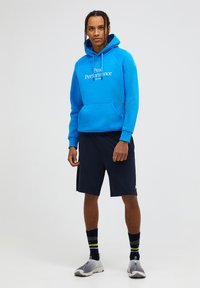 Felpa con cappuccio di un vivace blu con logo bianco, abbinata a pantaloni corti blu navy. Il modello indossa sneakers grigie e calze a righe multicolori. Design semplice.