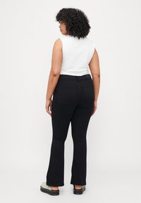 Jean noirs évasés avec une taille mi-haute, des poches arrière et une texture de tissu lisse. Associé à un haut sans manches blanc et à des chaussures vertes.