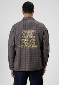 Geruit flannel overhemd in donkergrijs en zwart met geel geborduurde tekst op de achterkant. Voorzien van een klassieke kraag en lange mouwen.