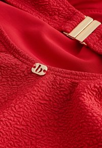 Primo piano di tessuto rosso testurizzato con una fibbia dorata e un piccolo dettaglio logo dorato su un capo di abbigliamento.