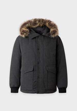 Jeune homme portant une veste d’hiver noire à capuche avec bordure en fourrure, un pull bleu zippé, un pantalon foncé et des mocassins noirs, debout avec une main dans la poche.